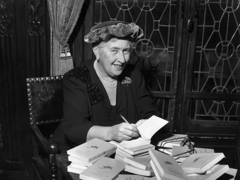 An older Agatha Christie sits at a table signing a large pile of her books